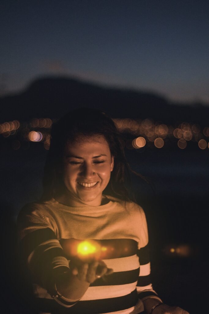 Smiling woman holding a lighted object outdoors at night with city lights bokeh in the background.
