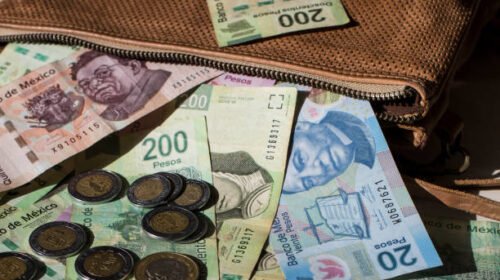 A mixture of Mexican pesos and coins are semi contained within a leather handbag. The bag is lying on a flat surface.