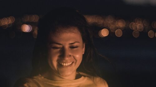 Smiling woman holding a lighted object outdoors at night with city lights bokeh in the background.